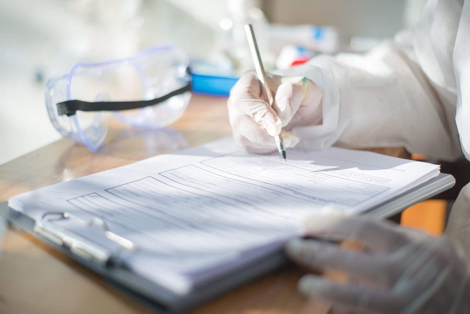 Gloved hands completing a batch record on a clipboard beside safety goggles