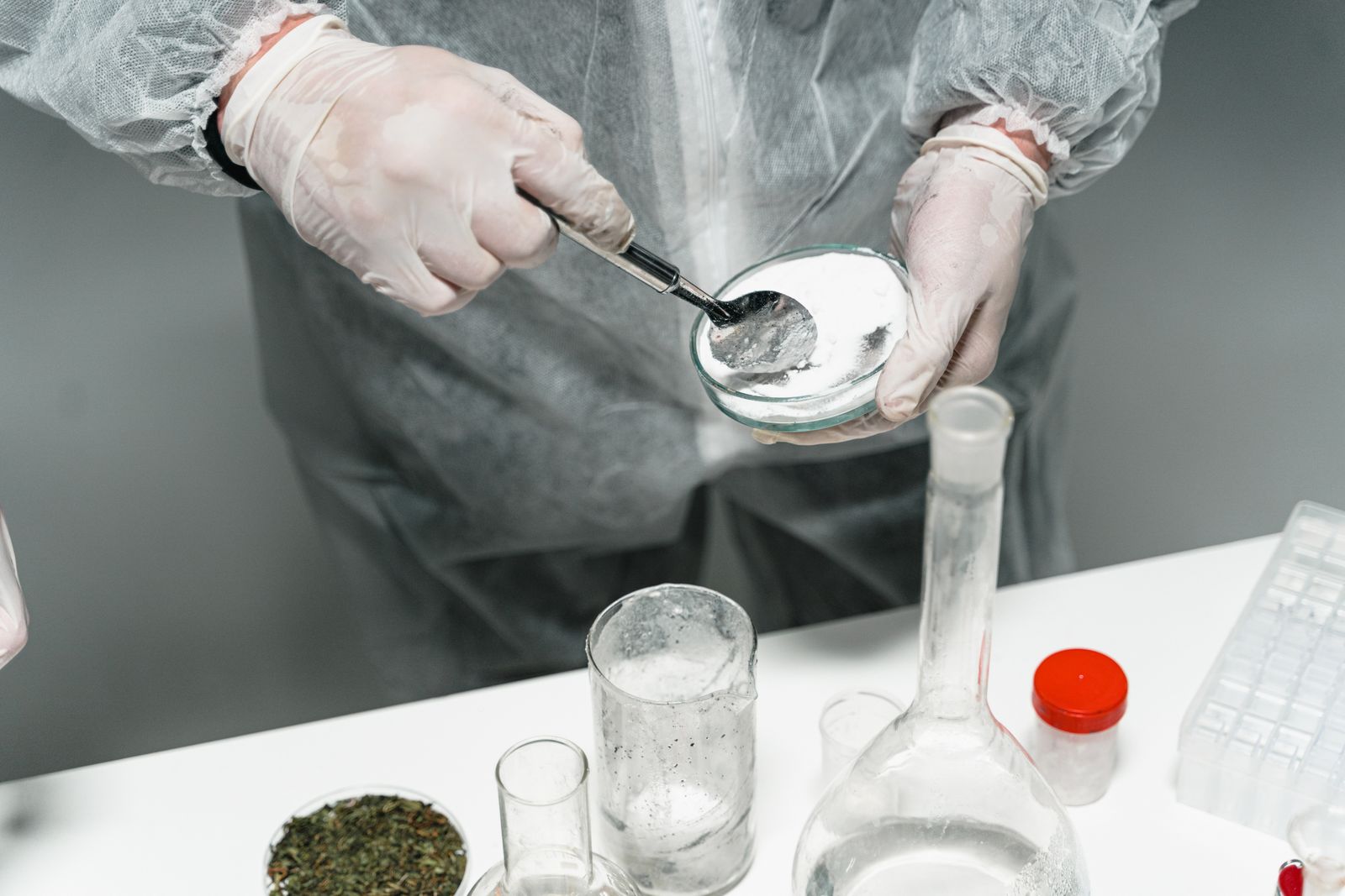 A technician in a coverall and gloves transferring a white crystalline powder onto a watch glass