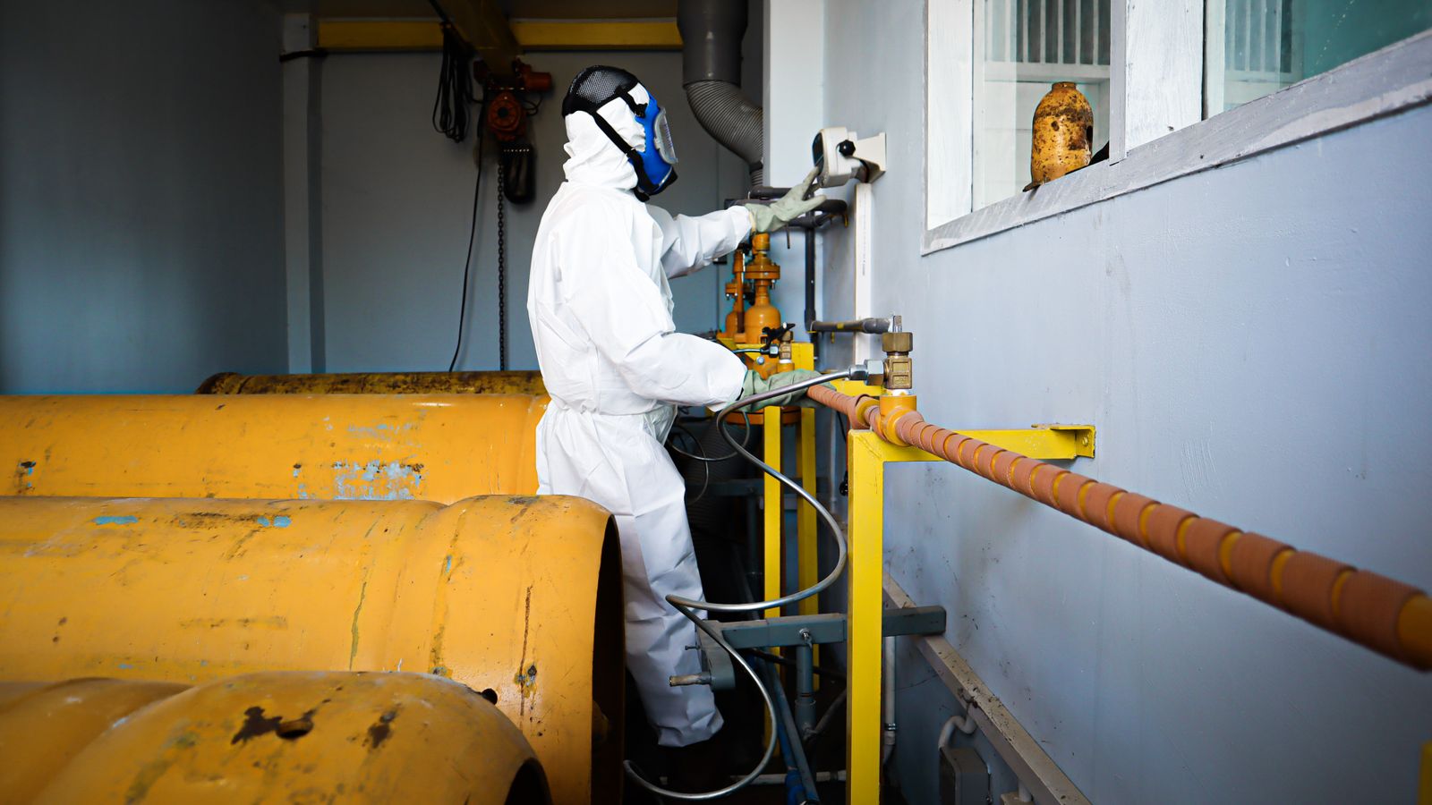 A worker in a full protective coverall, gloves and respirator operating plant equipment, face not visible